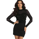 YNIQUE model wearing black holiday party sequin blouse with sheer long sleeves and fitted black mini skirt by Christmas tree