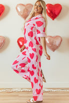 Woman wearing YNIQUE heart print valentine pajamas set with red hearts on pink fabric posing indoors