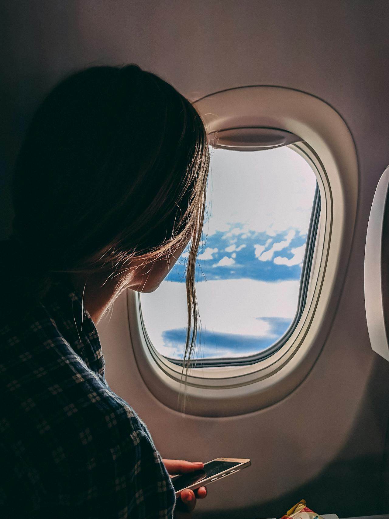 Woman looking out airplane window while using her phone with solo travel tips for women