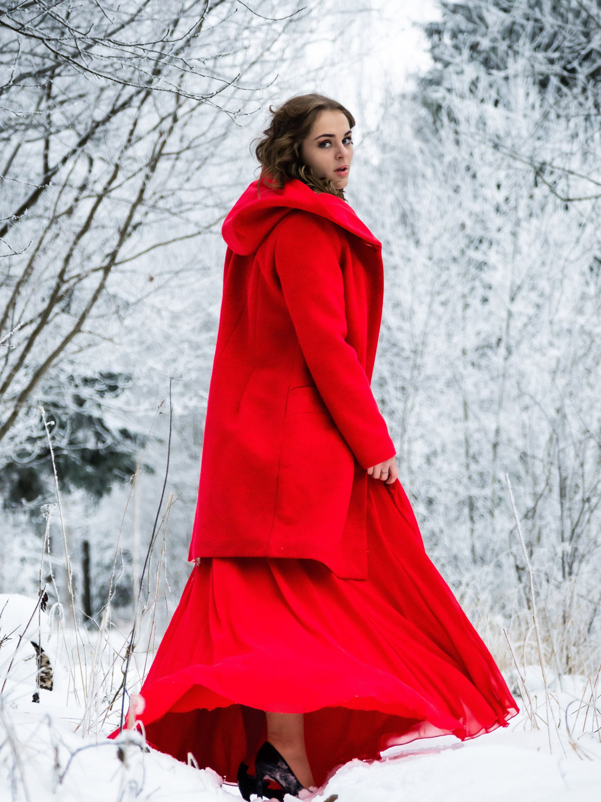 Woman in a dramatic red winter coat and gown standing in a snowy forest