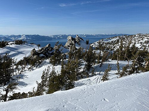 Snow-covered ski run with granite boulders and a view of Lake Tahoe in the distance