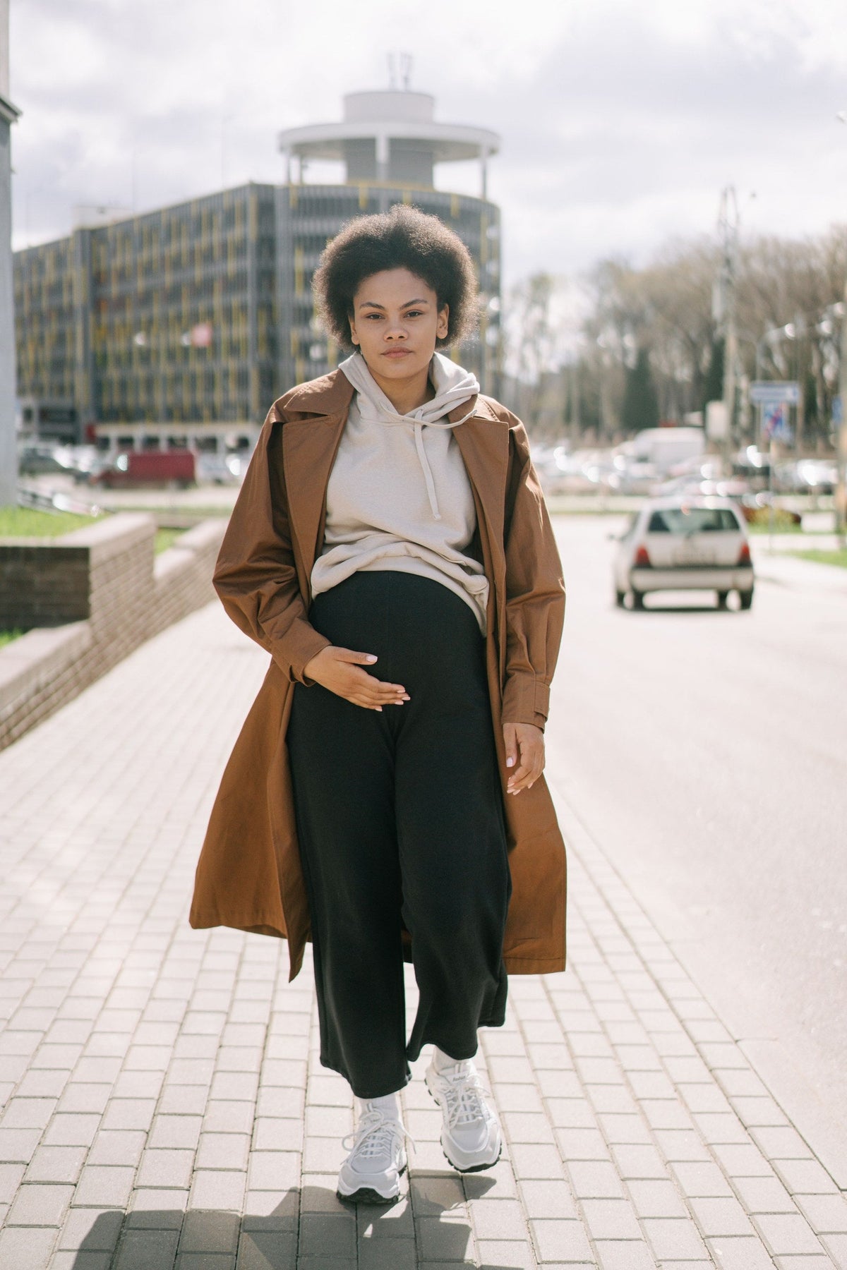 Pregnant woman in a trench coat, hoodie, and sneakers walking on a city sidewalk, dressed for travel