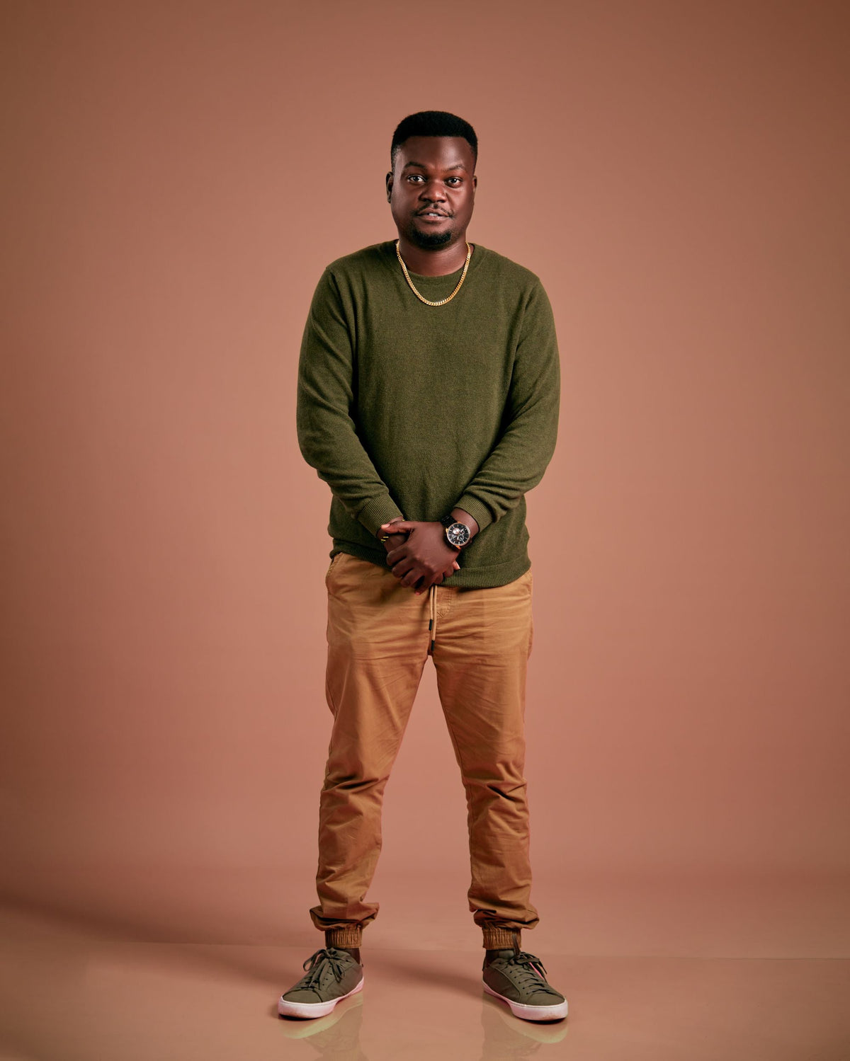 Man wearing a green sweater and tan travel pants standing in a studio portrait