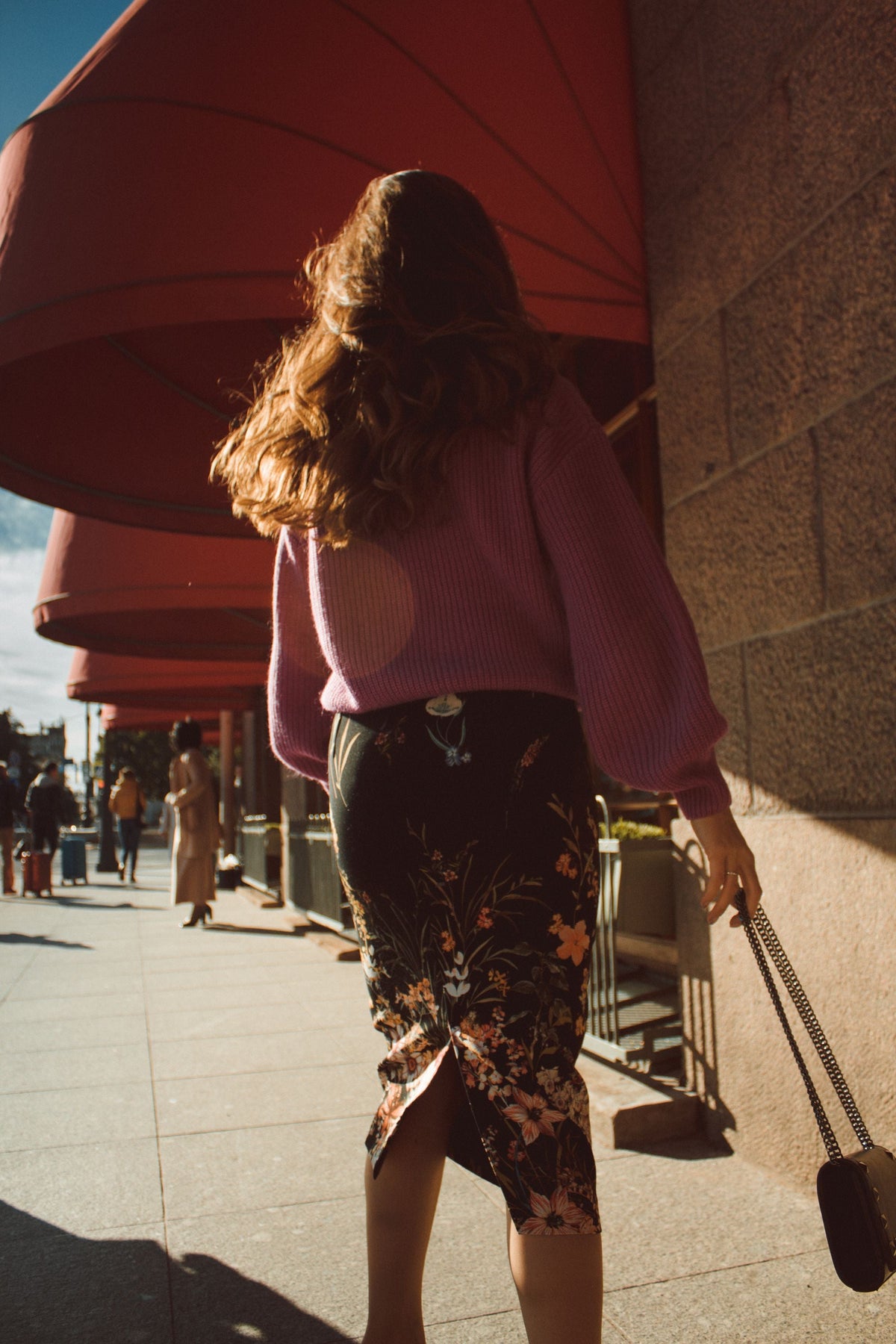 Woman wearing a pink sweater and floral skirt walking down a sunny city street