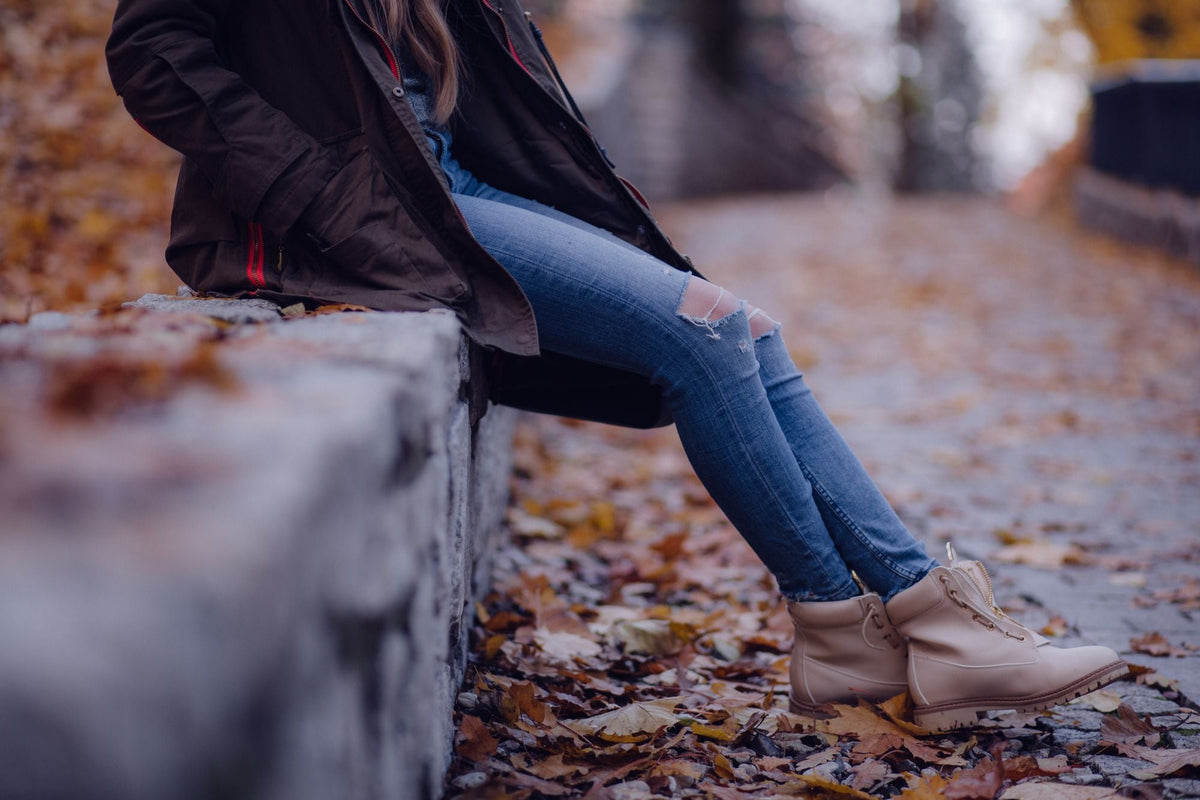 Woman sitting on stone wall wearing tan boots among autumn leaves