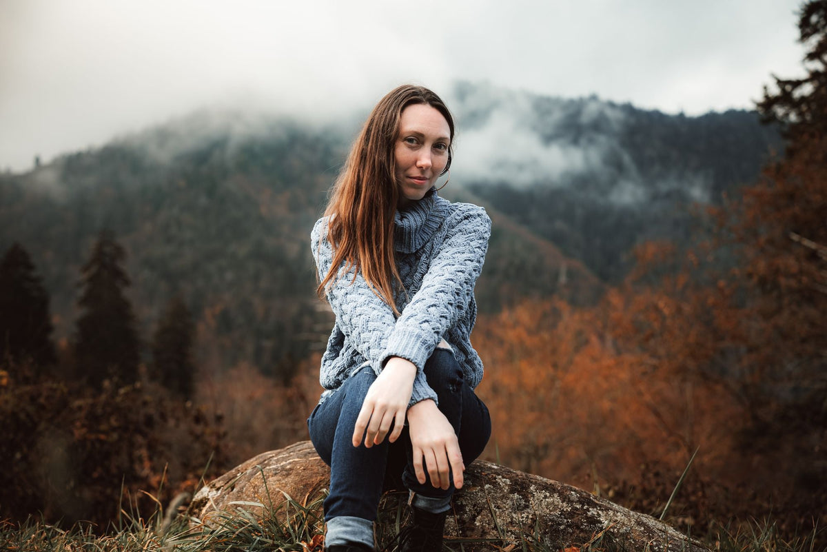 Woman wearing a cozy grey cable knit sweater sitting on a rock in a misty autumn mountain landscape