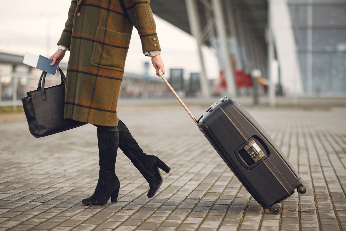 Woman in plaid coat and boots walking with suitcase and passport near airport terminal