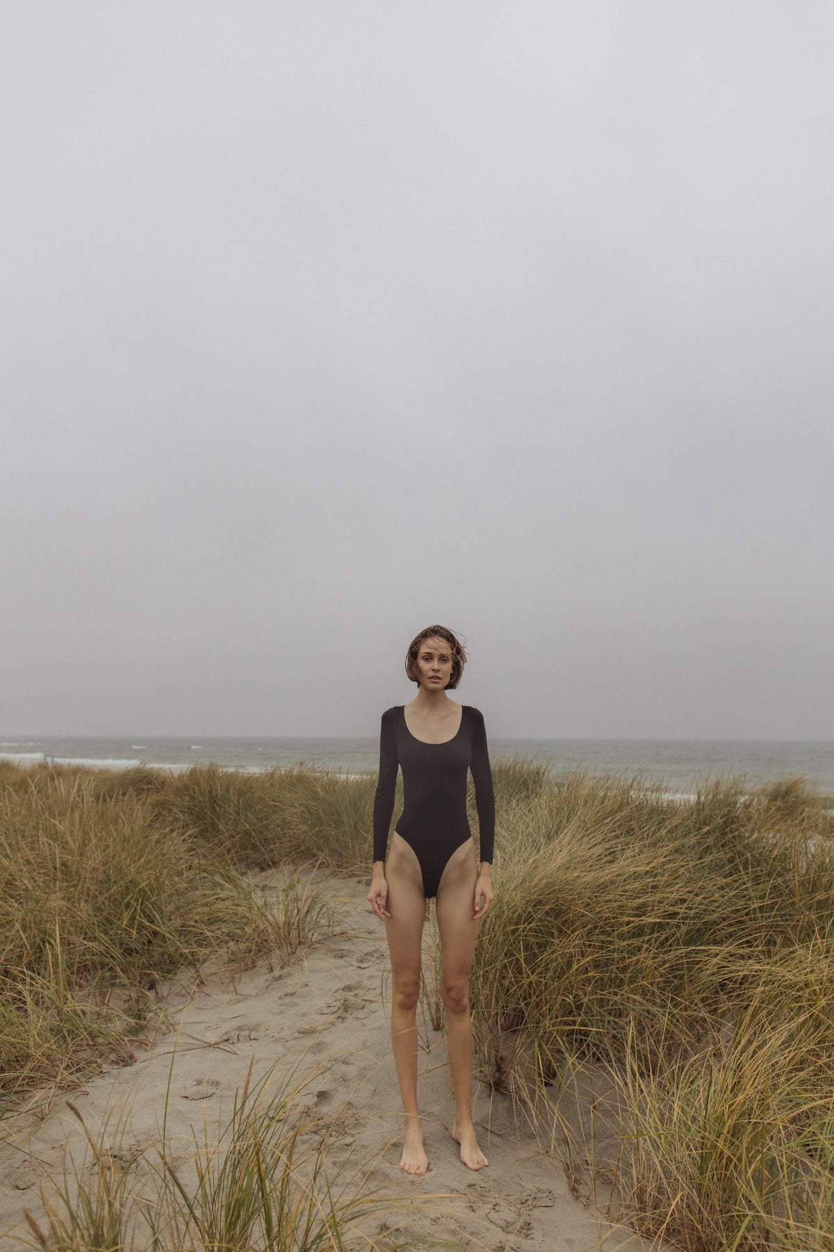 Woman in black one-piece swimsuit standing on grassy dune, overcast