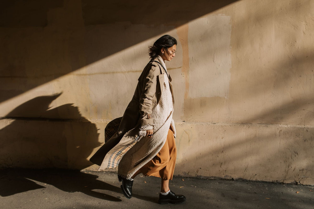 Woman walking in layered trench coat, sweater, and skirt in autumn light