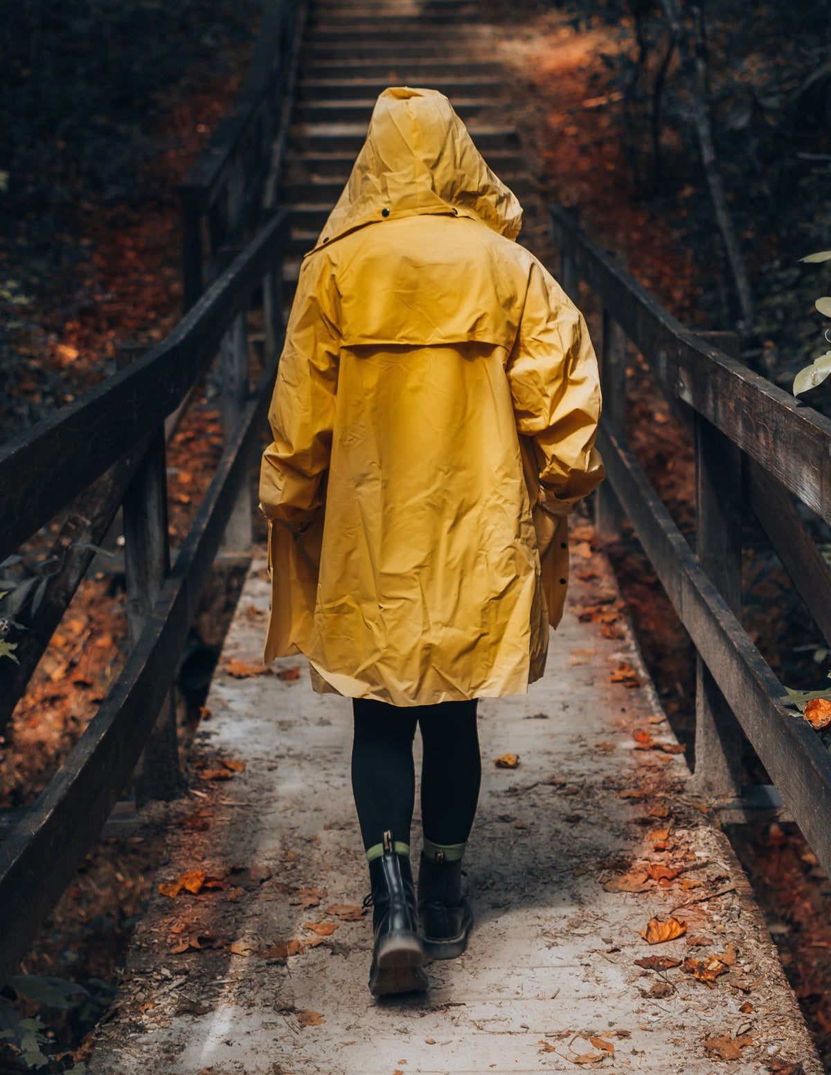 Person in a yellow hooded rain jacket walking away down a wooden forest path toward stairs in autumn