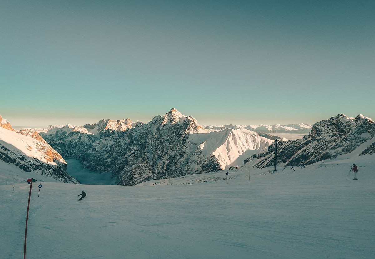 Skiers on a snowy alpine slope at a ski resort with dramatic snow-capped mountain peaks in the background