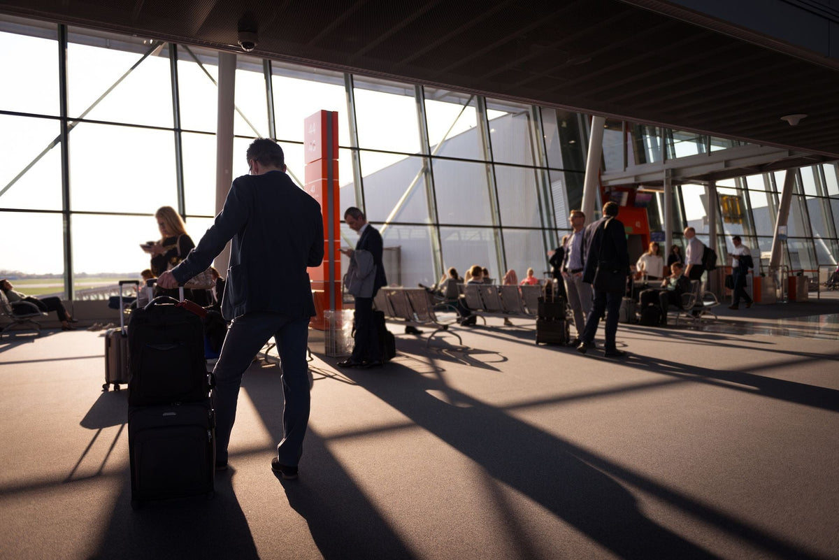 Traveler pulling luggage through a busy, sunlit airport terminal.