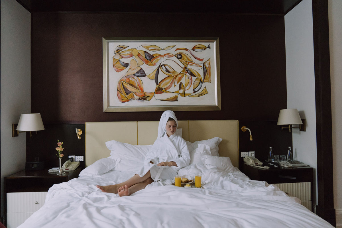 Woman in a white robe relaxing on a hotel bed with breakfast