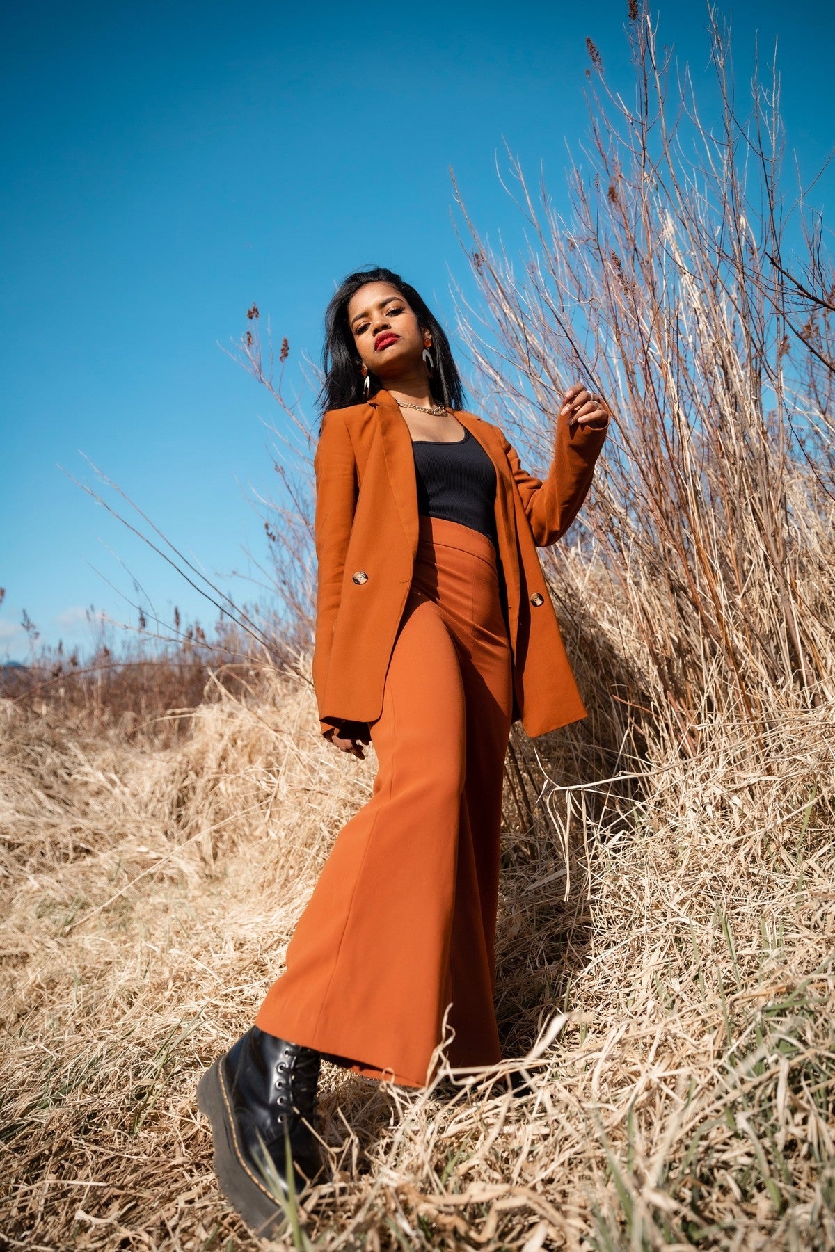Woman wearing a rust orange tailored blazer and matching trousers standing in a dry grass field under a blue sky