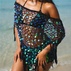 A woman on a beach wearing a black off-the-shoulder mesh cover-up with iridescent blue, green, and teal sequins and fringe.