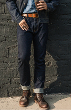 Person wearing a denim jacket, jeans, and brown boots against a black brick wall.