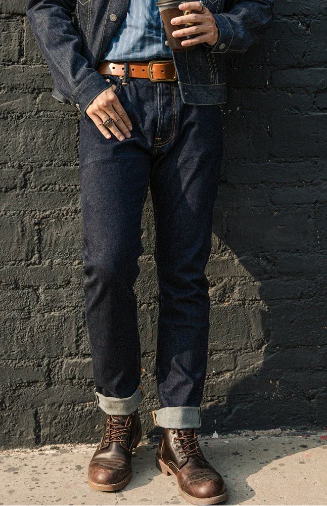 Person wearing a denim jacket, jeans, and brown boots against a black brick wall.