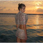 A woman stands at the edge of the ocean at sunset wearing a sparkling silver sequined two-piece outfit with a short puff sleeve top and matching mini skirt.