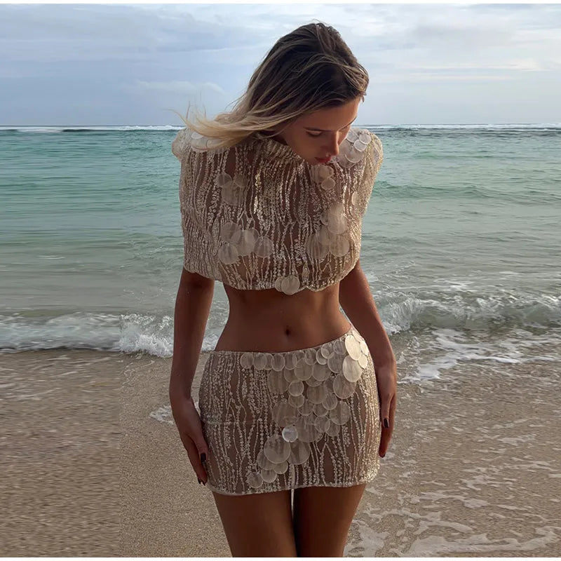 A woman stands on a beach wearing a beige textured two-piece outfit with a cropped top and matching mini skirt, featuring a scalloped shell-like pattern.