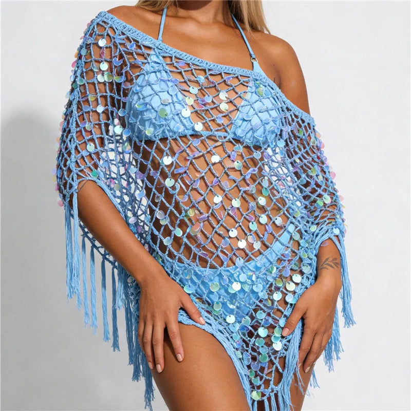 A woman wearing a light blue crochet bikini cover-up with fringe and iridescent sequins, shown against a plain light gray studio background.