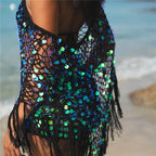 A black open-knit fringed cover-up with iridescent blue, green, and purple sequins, worn by a model on a beach with the ocean in the background.