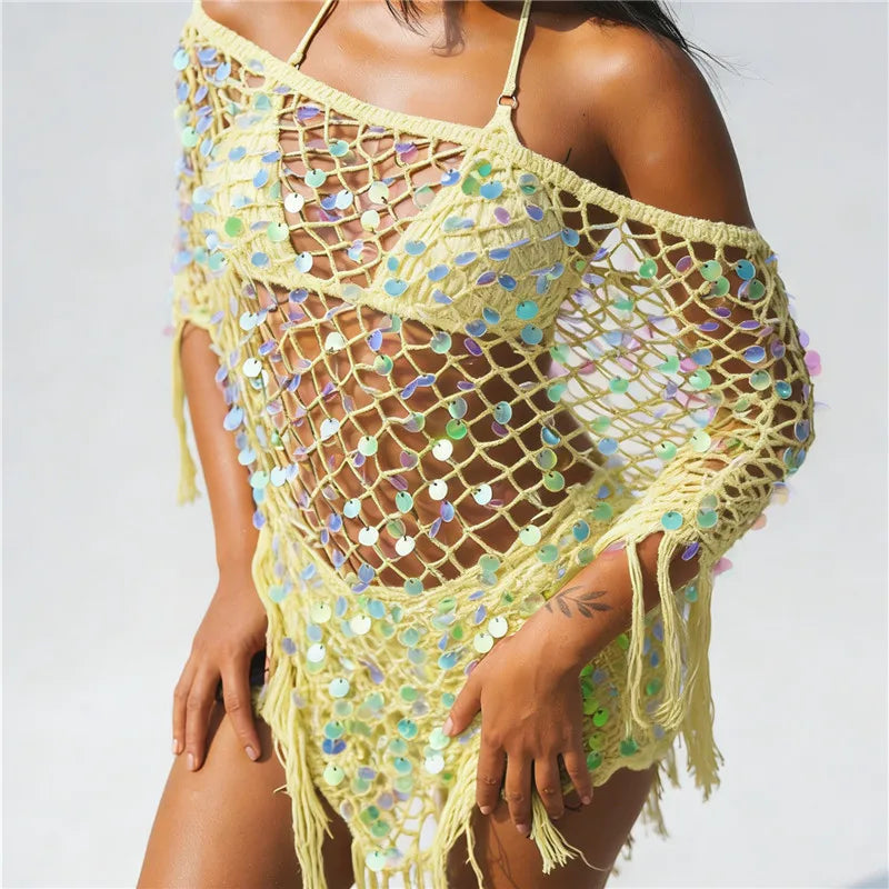 A woman wearing a pale yellow crochet beach cover-up with iridescent sequins and fringe, shown against a plain light background.