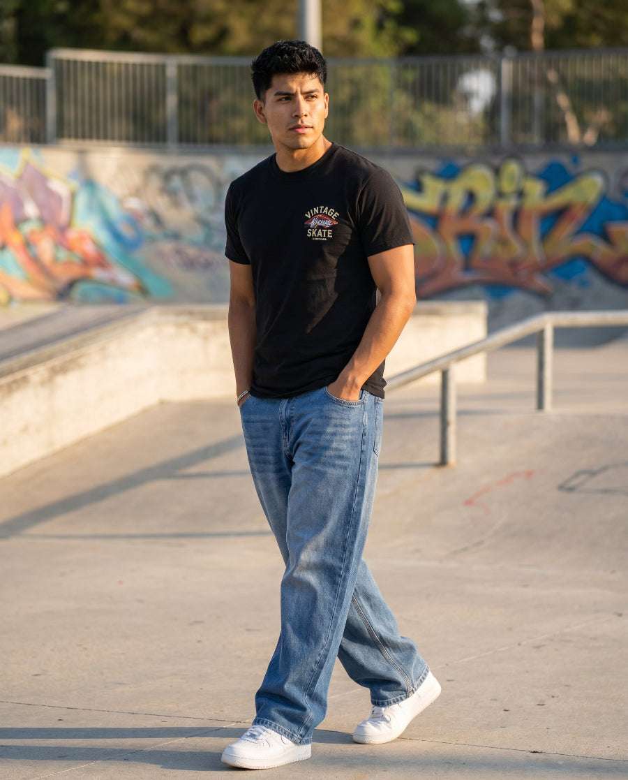 Man wearing medium blue wide-leg baggy jeans walking through skate park at golden hour