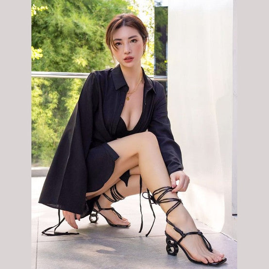 Black strappy vacation sandals by Rag Company worn by woman in black dress sitting outdoors