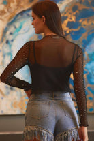 Model back view wearing YNIQUE black rhinestone mesh top with sheer long sleeves and fringe denim shorts