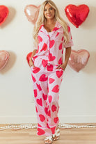 Woman wearing heart print valentine pajamas set with red hearts on pink fabric standing indoors with heart-shaped balloons