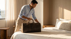 Man placing vintage leather weekender bag on hotel bed, cinematic landscape view with warm morning light