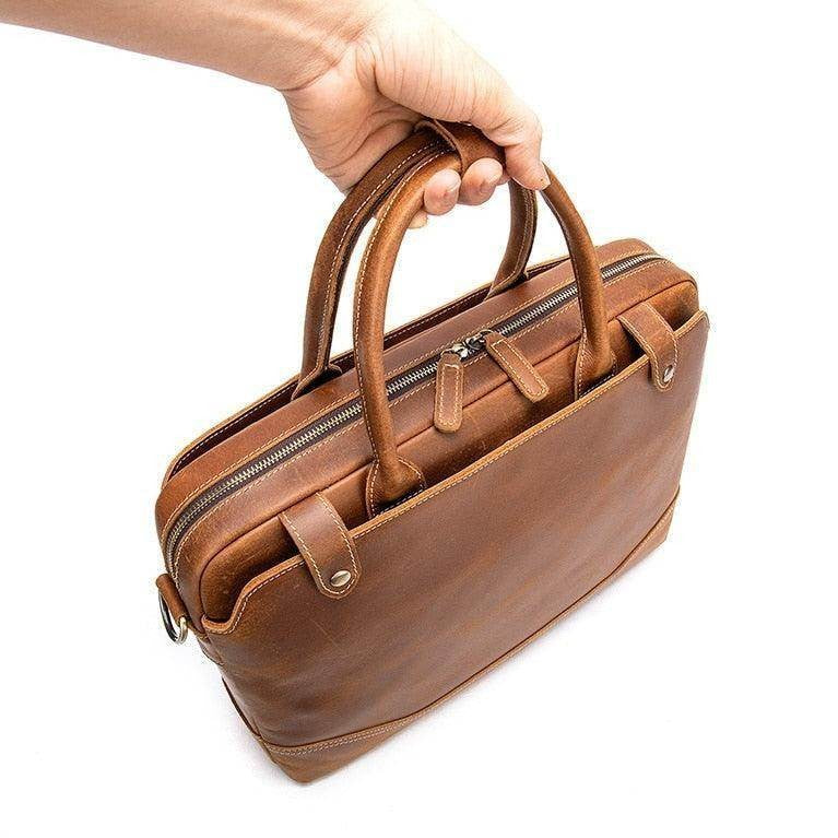 Hand holding a brown mens leather satchel bag with dual handles and zippered compartments