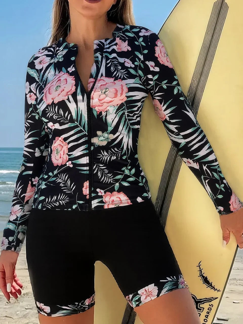VacationGrabs long sleeve floral zip-up surfing swimsuit set with black shorts on a beach with a surfboard