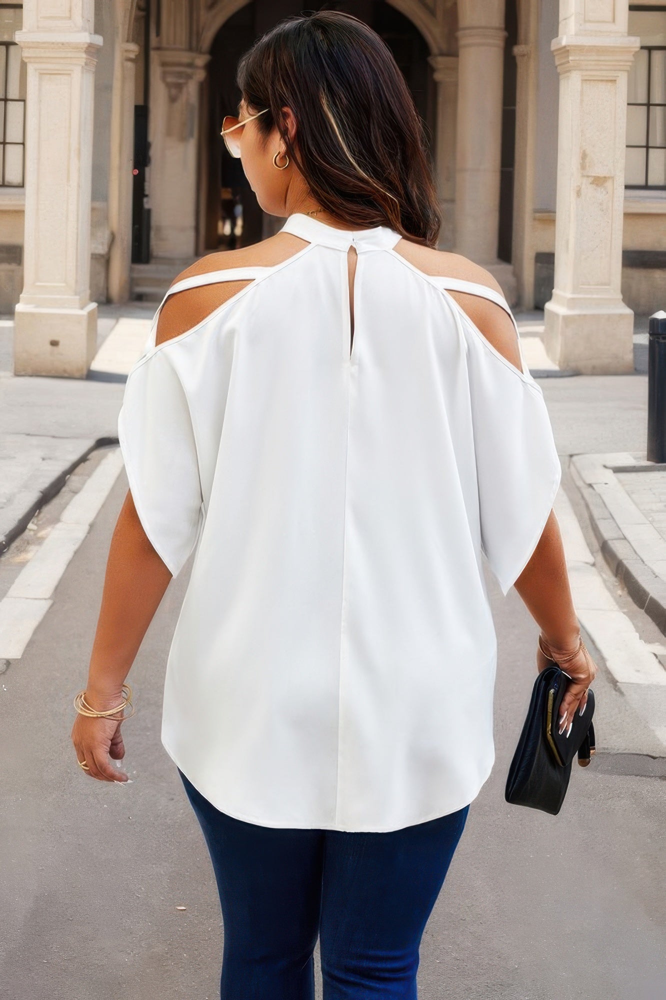 Back view of trendy plus size cold shoulder shirt in white worn by woman with dark hair holding black clutch